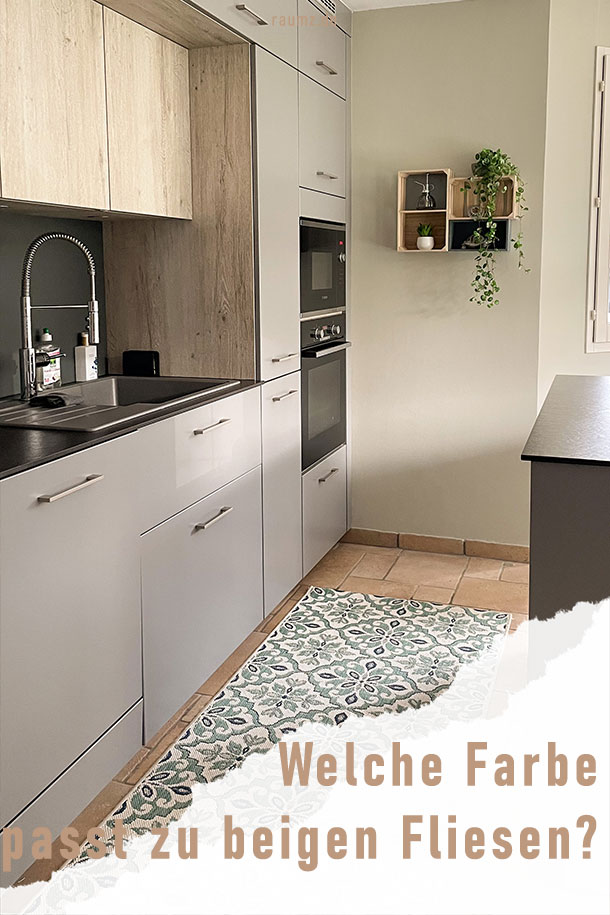 Ein hochformatiges Bild einer glänzend grauen Küche, silberner Amatur und schwarzer Arbeitsfläche. Gerahmt wird die Küche von Birkenholz akzenten, saleigrünen Wänden und beigen Fliesen. Auf den Fliesen befindet sich ein salbeigrüner Teppich mit Muster. Das Bild ist an der rechten Ecke gerissen, wo sich der Titel befindet: Welche Farbe passt zu beigen Fliesen?