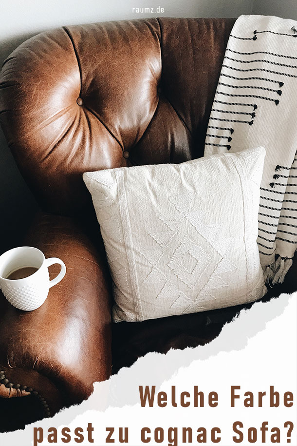 Hochformatiges Bild über das Thema "Welche Farbe passt zu cognac Sofa?". Zu sehen ist ein cognacfarbener Sessel mit weiß-gemustertem Kissen und Decke, sowie einer weißen Tasse. Das Bild ist in der unteren Ecke eingerissen und zeigt den Titel.