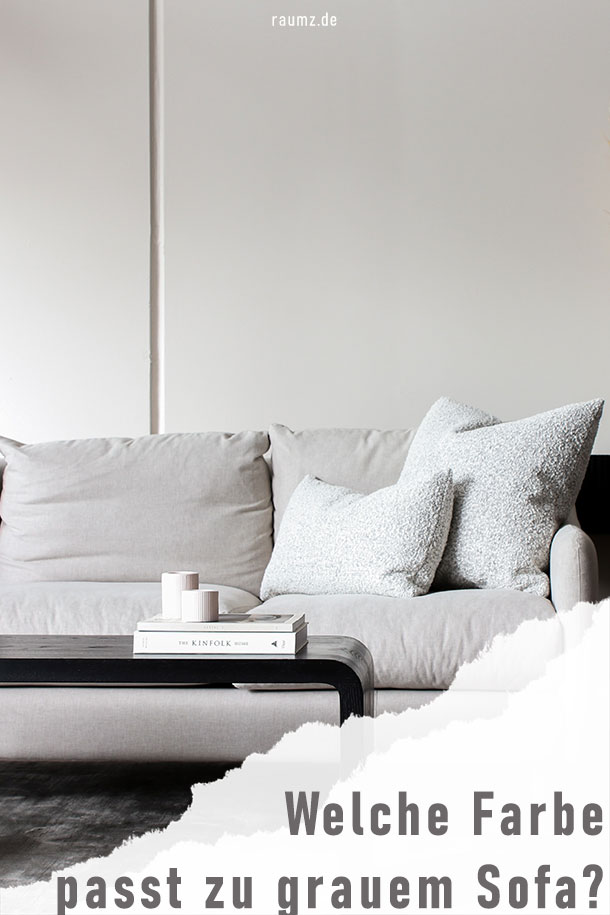Hochformatiges Bild über das Thema "Welche Farbe passt zu cognac Sofa?". Zu sehen ist ein graues Sofa mit hellgrauen Kissen und Wandfarbe. Vor dem Sofa erkennt man einen schwarzen Beistelltisch mit weißem Büchern und Kerzen. An der unteren rechten Ecke ist das Bild eingerissen und zeigt den Titel des Beitrags "Welche Farbe passt zu grauem Sofa".