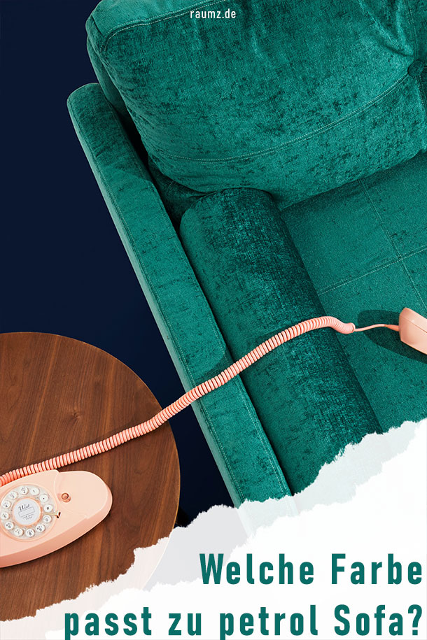 Hochformatiges Bild zu dem Beitrag "Welche Farbe passt zu petrol Sofa?". Zu sehen ist ein petrolfarbenes Sofa aus Samt. Das Sofa steht auf einem dunkelblauen Teppich. Links neben dem Sofa befindet sich ein dunkler Holztisch mit einem rosé Telefon.