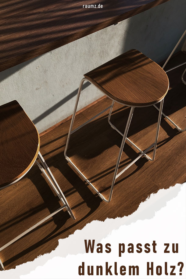 Hochformatiges Bild mit einem Ausschnitt einer Küche aus dunklem Holz. Zu sehen sind drei Stühl im Barhocker-Stil. So bestehen Sie aus einer Sitzplatte aus dunklem Holz und weißen Metallfüßen. Sie stehen von einem Esstisch. Dieser Esstisch ist in die Wand eingelassen. Die Wand selbst ist in Betonoptik gehalten. An der unteren rechten Ecke ist das Bild eingerissen und man erkennt den Titel: Was passt zu dunklem Holz?
